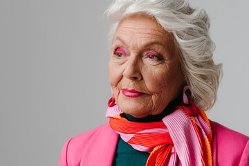 Elegant elderly woman with make-up wearing fashionable clothing and radiating self-love on grey background