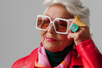 Fashionable senior woman in red leather jacket adjusting trendy eyeglasses on grey background