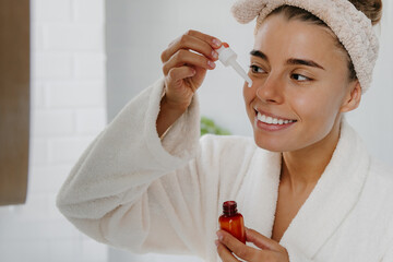 Happy young woman in bathrobe applying cosmetic serum on face while standing in bathroom