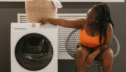 Young African woman select settings for laundry while sitting in comfortable chair near the washing machine - 765793743