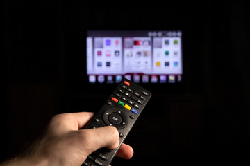 A person switches channels on a TV using a remote control. In the hand of a person, a TV remote control, a close-up photo, against the background of a blurred TV