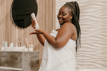 Happy plus size African woman covered in towel applying cosmetic cream on hand in bathroom