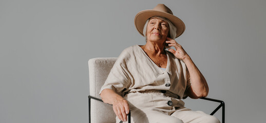 Elegant gray hair senior woman in hat relaxing in comfortable chair at the sunny studio