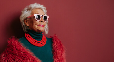 Fashionable elderly woman in stylish eyeglasses looking cool and trendy while standing on red background