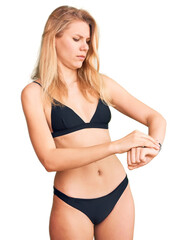 Obraz premium Young beautiful blonde woman wearing bikini checking the time on wrist watch, relaxed and confident