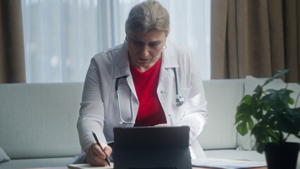 Senior female doctor in white coat work at workplace sit on sofa, writes notes, prepare report, filling patient card, prescribe medications	
 - Powered by Adobe