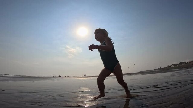 Child runs cheerfully through shallow water along beach, illuminated by rays setting sun. Sun paints in warm shades child. Sun plays with reflections on water, emphasizing joyful moments childhood.