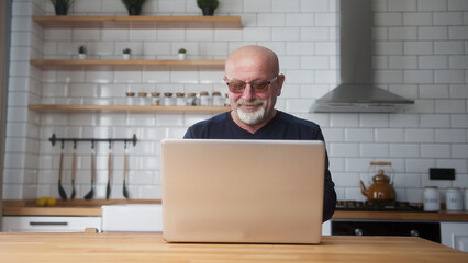 Smiling old man in glasses sit in kitchen using laptop, spend free time on internet, enjoy communication in social media, resting at home. Wireless, modern tech, e-shopping	
