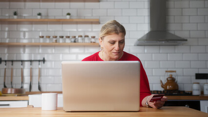 Middle aged woman sittin in the kitchen enter credit card number on laptop for shopping online,...