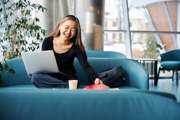 Young laughing asian female IT employee taking notebook while working on laptop