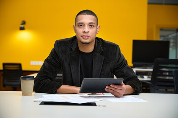Fototapeta premium Serious young male employee sitting at table and holding tablet