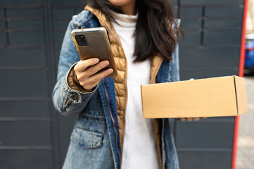 Young woman using smart phone while standing with a parcel delivered with post office machine with...
