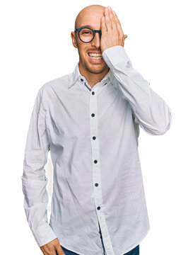 Bald man with beard wearing business shirt and glasses covering one eye with hand, confident smile on face and surprise emotion.