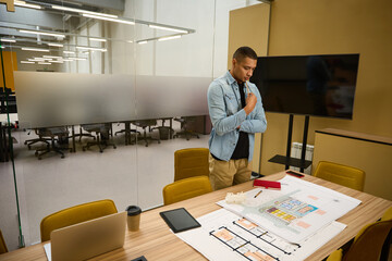Thoughtful young employee standing near table and looking at plan