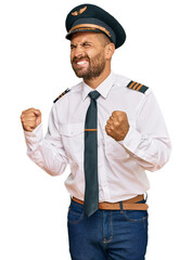 Handsome man with beard wearing airplane pilot uniform excited for success with arms raised and...