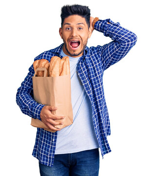 Handsome latin american young man holding paper bag with bread crazy and scared with hands on head, afraid and surprised of shock with open mouth