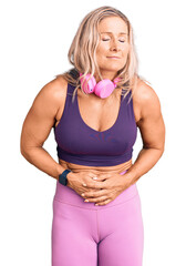 Middle age fit blonde woman wearing gym clothes and using headphones with hand on stomach because...