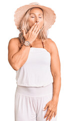 Middle age fit blonde woman wearing summer hat bored yawning tired covering mouth with hand. restless and sleepiness.