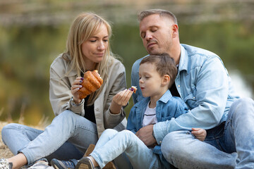 Caucasian mother, father and daughter are on a picnic in a forest on a lake background during fall,...