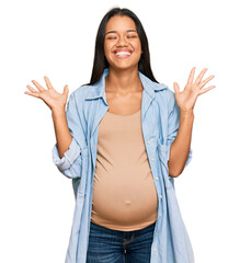 Beautiful hispanic woman expecting a baby showing pregnant belly celebrating mad and crazy for...