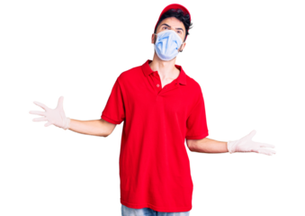 Young hispanic man wearing delivery uniform and medical mask crazy and mad shouting and yelling with aggressive expression and arms raised. frustration concept.
