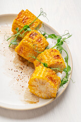Closeup view of grilled slices of yellow corn or maize decorated with fresh pea microgreens with sauce served on plate on white wooden table prepared for healthy vegetarian appetizer as mexican food