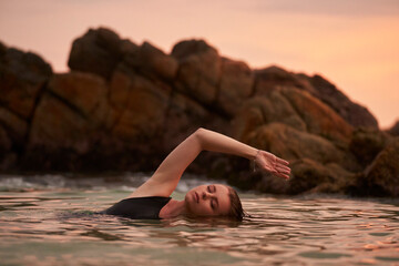 Elegant woman relaxes in sea at sunset, immersed in water near rocks. Female enjoys tranquil moment, healthy ocean swim. Natural wellness, peaceful retreat, harmonious lifestyle, mind-body balance.
