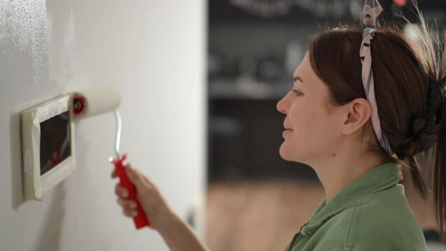 Slow motion. Close-up. A smiling woman carefully paints the corridor wall with a wide paint roller with white paint and talks