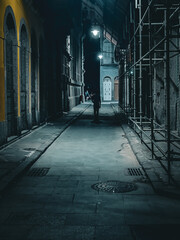 Rua estreita à noite com pouca iluminação e prédios antigos com um homem caminhando. - obrazy, fototapety, plakaty