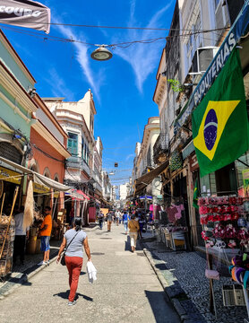 Rua comercial com bastante movimento de pessoas, muitas lojas e uma bandeira do Brasil pendurada em um dia de muito sol com c&eacute;u azul.