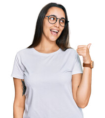 Young hispanic woman wearing casual white t shirt smiling with happy face looking and pointing to the side with thumb up.