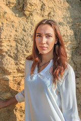 A beautiful girl in a white shirt sits and stands against the background of a large stone on the beach
