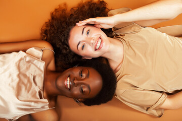 two pretty girls african and caucasian blond posing cheerful together on brown background,...