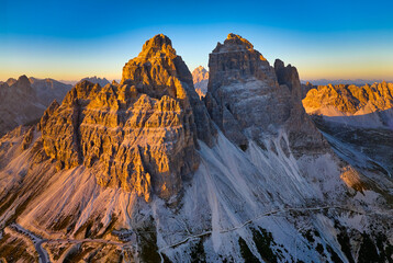Aerial view from high altitude of Tre Cime di Lavaredo peaks orange lit by the setting sun, with a Dolomites mountain panorama in the background, blue sky with a yellow horizon band. Italy