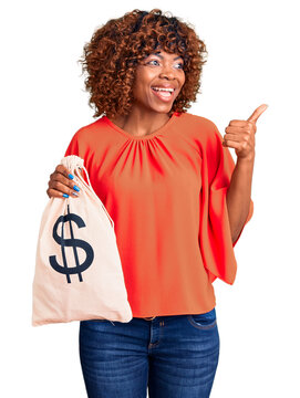 Young african american woman holding money bag with dollar symbol pointing thumb up to the side smiling happy with open mouth