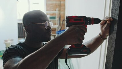 African American man in stained t-shirt and glasses drilling screw into door frame when doing home renovation or repairment
