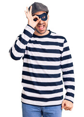 Young handsome man wearing burglar mask doing ok gesture with hand smiling, eye looking through fingers with happy face.