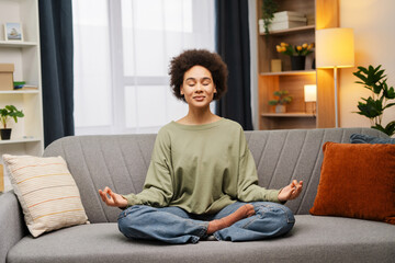 Beautiful, African American woman in casual clothes sitting on comfortable sofa in lotus position