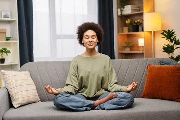 Tuinposter Beautiful, African American woman in casual clothes sitting on comfortable sofa in lotus position © Maria Vitkovska