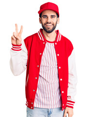 Young handsome man with beard wearing baseball jacket and cap smiling with happy face winking at...