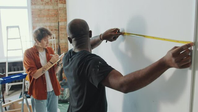 African American man taking wall measurements with tape measure and telling them to young Caucasian wife writing down notes in copybook when doing home renovation together