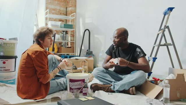 Young Caucasian wife and African American husband sitting on the floor in living room among boxes and tools, examining paint samples and sharing ideas about home renovation. Zoom Shot