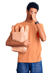 Young african american man holding paper bag with bread smelling something stinky and disgusting,...