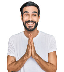 Young hispanic man wearing casual white t shirt praying with hands together asking for forgiveness smiling confident.