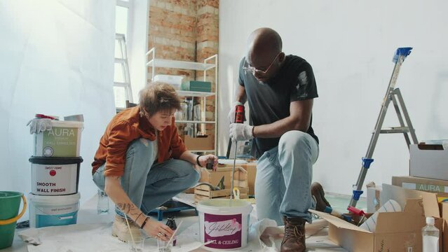 Black man stirring paint with drill as his Caucasian wife adding liquid colourant to bucket and checking color before painting walls during home renovation. Zoom Shot