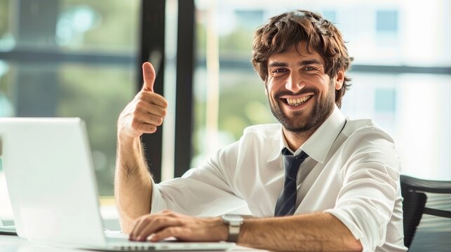 A good-looking man is happily using a laptop and showing a thumbs up gesture with a big smile on his