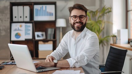 Portrait of young happy attractive bearded business man in white shirt and glasses sitting at the desk of his workplace with laptop with charts at the office, looking cheerful at camera and smiling