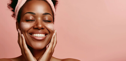 A cheerful African American woman with a headband smiles brightly while applying a creamy skincare product to her cheek