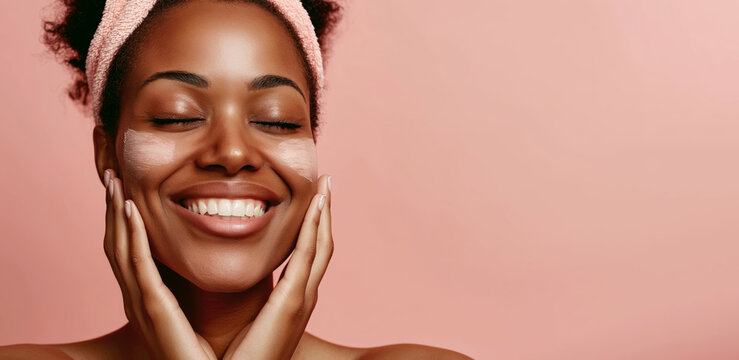 A cheerful African American woman with a headband smiles brightly while applying a creamy skincare product to her cheek