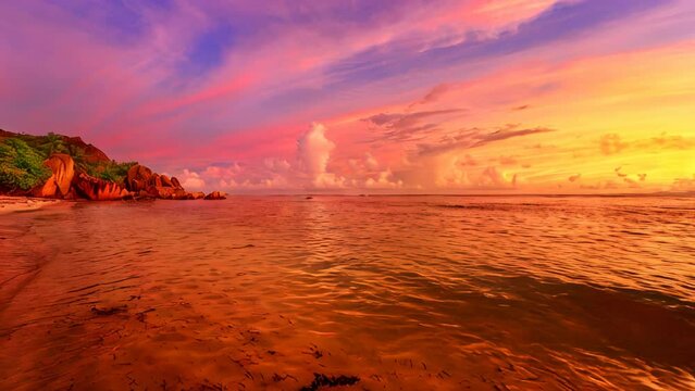 Seychelles, La Digue, Anse Source d'Argent at sunset. A picturesque scene with a sky painted in vibrant hues, adorned by granite rock formations and palm trees. The twilight sun casts its warm glow.